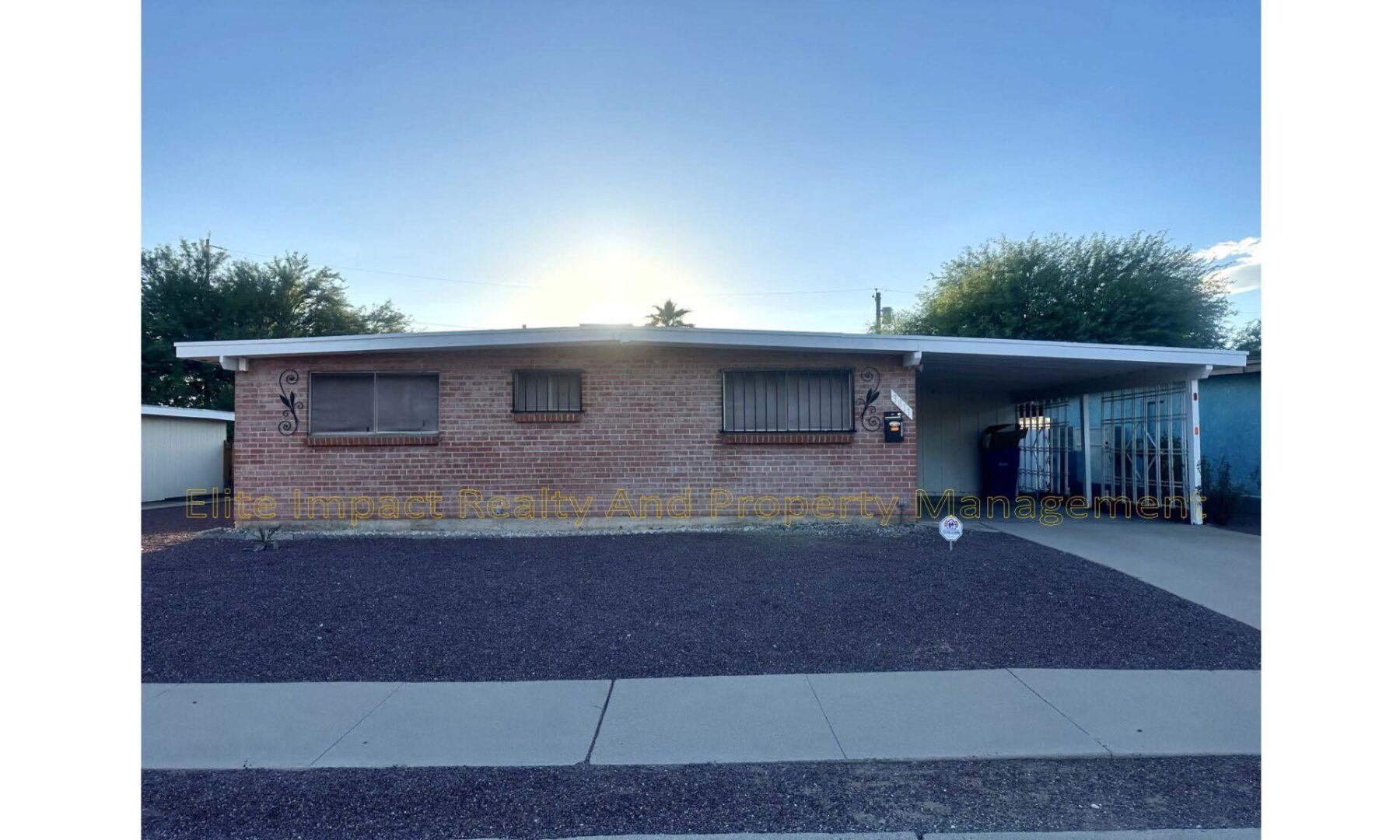 Single-story brick house with a carport and gravel yard at sunset.